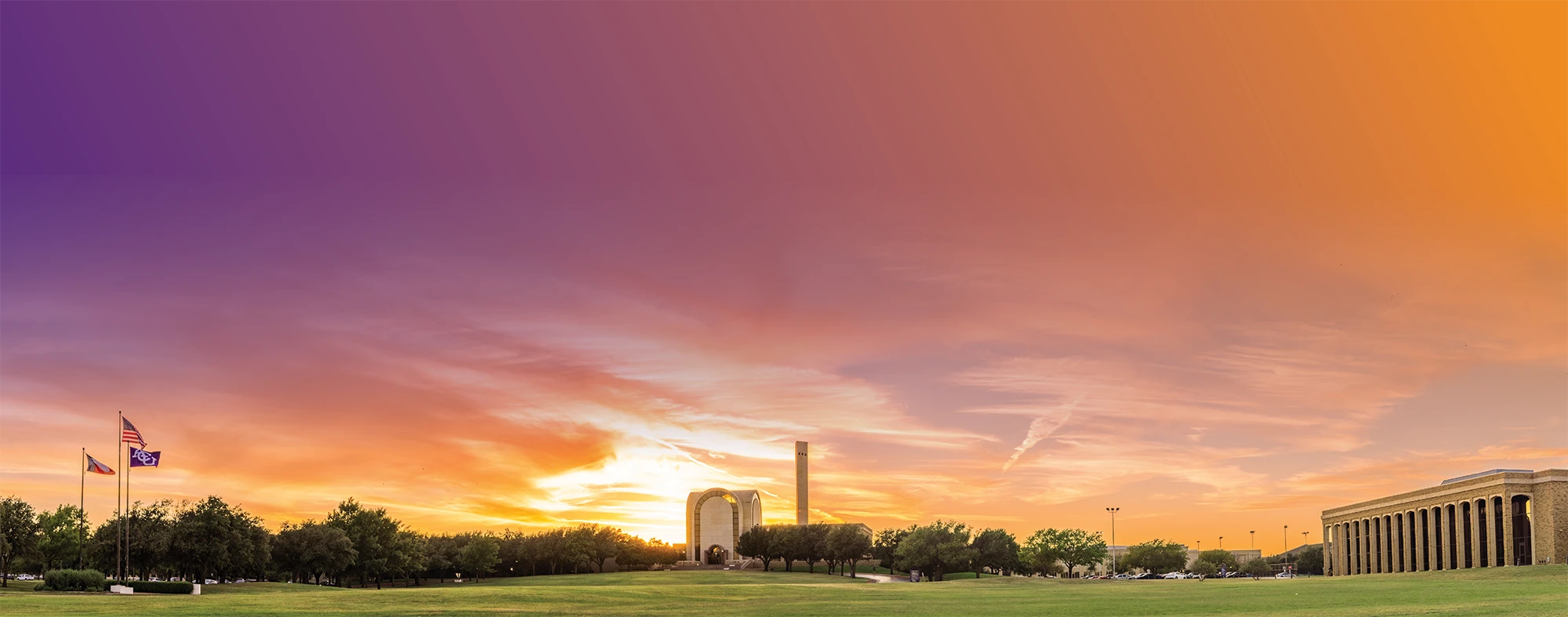 Sunset view of the Abilene Christian University campus featuring a wide lawn, campus buildings, and three flags—Texas, American, and ACU’s flag—under a vibrant purple-to-orange sky.