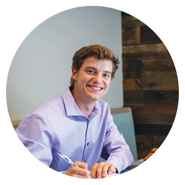 Portrait of a smiling male student in a lavender dress shirt, seated indoors with a pen and notebook, in front of a wood-paneled wall.