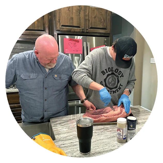 Older man and younger student preparing a large cut of meat together in a kitchen; the student wears an ACU Biology Department sweatshirt and gloves while seasoning the meat.