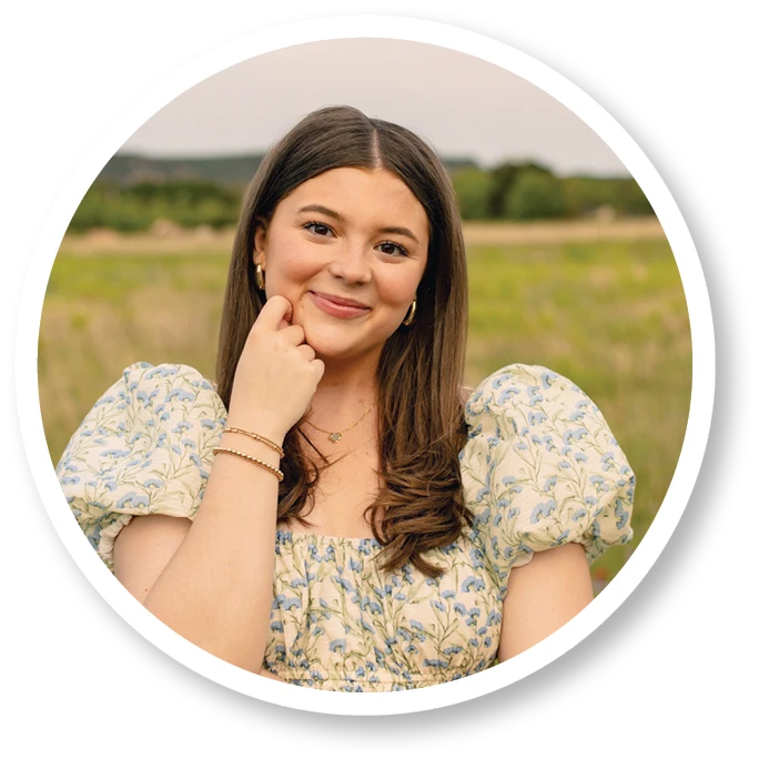Portrait of a smiling young woman with long dark hair wearing a floral puff-sleeve dress, posing with her hand resting on her chin in a grassy outdoor setting. Circular white and black border surrounds the image.