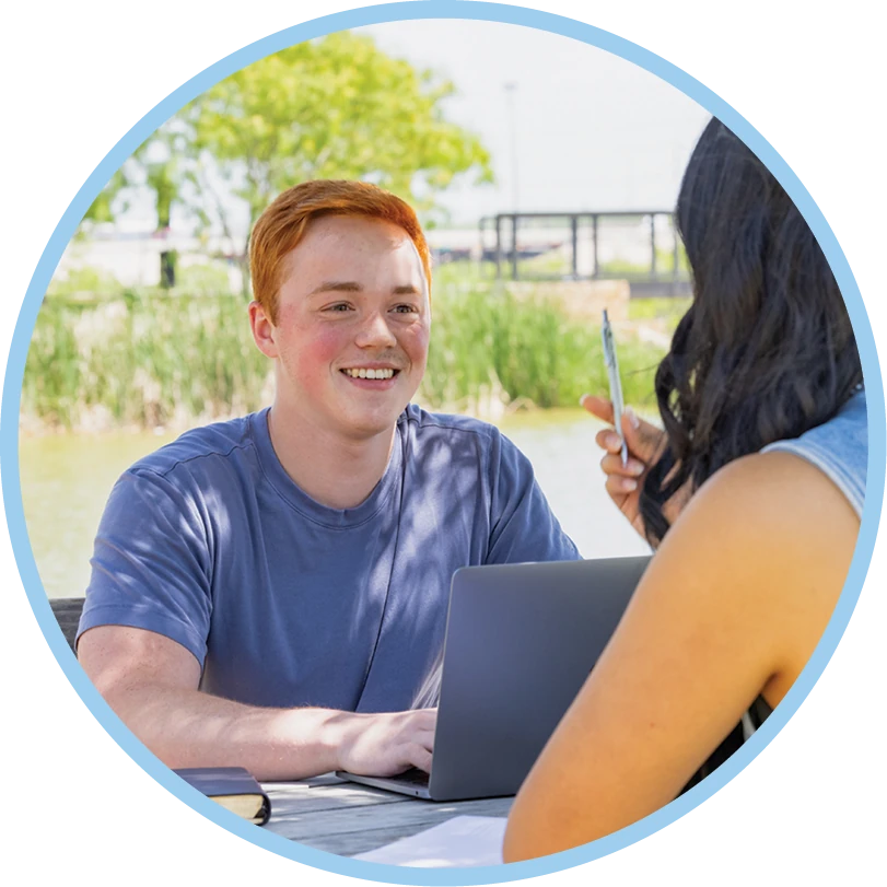 ACU student talks with another student while using a laptop outdoors.