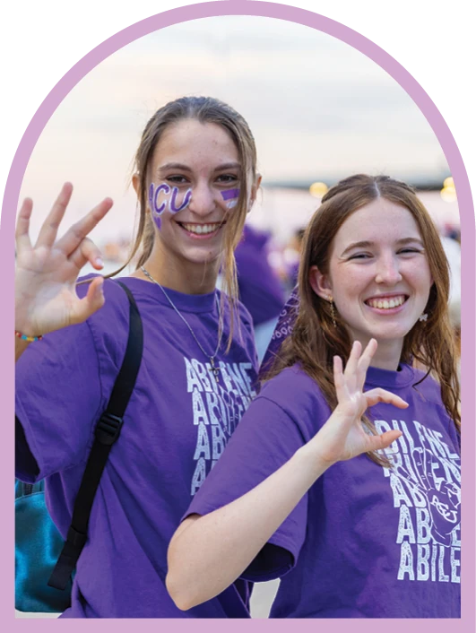 Two ACU students in purple shirts smiling and making the W-C hand sign.